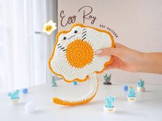 a hand is holding an orange and white crocheted animal with cactus decorations around it