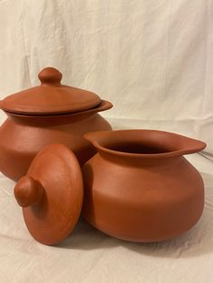 three clay pots with lids are sitting on a white cloth covered tablecloth in front of a white backdrop