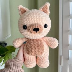 a hand holding a pink crocheted teddy bear in front of a green plant