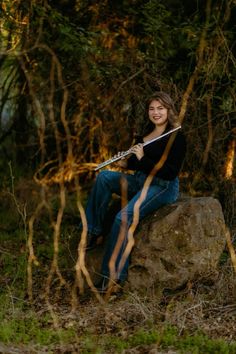 a woman sitting on top of a rock with a flute in her hand and trees behind her