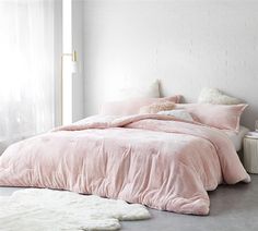 a bed with pink comforter and pillows in a white room next to a window