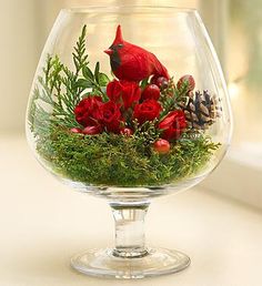 a red bird sitting on top of a glass bowl filled with flowers and greenery