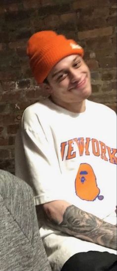 a young man wearing an orange beanie smiles at the camera while sitting in front of a brick wall