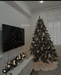 a decorated christmas tree in the corner of a living room with lights on it and a large tv mounted to the wall