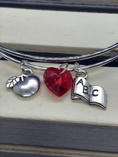 This bangle bracelet is a perfect gift for teacher in your life.  A small token of appreciation for all their sacrifices for our kids. 🔹 This bangle is made of steel. This features an apple charm, a book charm, and a red Swarovski crystal heart. All charms feature an antique finish.  🔹 The bangle comes packaged in an organza bag perfect for gift giving.   🔹 Gift options.  This item can be shipped to a gift recipient with a gift note if desired.  Please send me a message if this is a need you have and I will be happy to accomodate.  🔹 Customization options.  This bracelet can be customized with an initial charm at no extra cost. Just message me with the initial you would like to have.    🔹Please visit my shop for more of my crafty creations.    www.itsjustmyhobbyshop.etsy.com 🔹 If you Personalized Red Charm Bracelet As Gift, Nickel Free Charm Bangle For Personalized Gift, Nickel-free Bangle Charm Bracelet For Personalized Gift, Stackable Metal Charm Bracelet For Gift, Stackable Metal Charm Bracelet Gift, Metal Stackable Charm Bracelet Gift, Personalized Red Stainless Steel Jewelry, Red Adjustable Jewelry For Personalized Gift, Adjustable Red Jewelry For Personalized Gifts