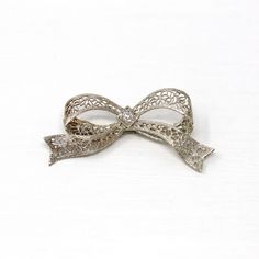 Gorgeous vintage circa 1930s era 14k white gold diamond bow brooch! This beautiful piece showcases a sparkly old European cut diamond center weighing an estimated .07 carats. The setting features ornate flower filigree with milgrain details. The brooch secures with a rotating c-clasp. An outstanding Art Deco Era piece of fine jewelry! *Sale - price reduced from $340 USD to $330 USD.  ERA - Circa 1930s - Art Deco  METAL / MATERIAL - 14k white gold, 1 old European cut diamond (estimated .07 cts, SI2, near colorless)  MARKINGS / HISTORY - Brooch clasp marked "14k"  CONDITION - Good antique condition. White gold metal has been professionally polished & cleaned. Very light age appropriate patina & wear remains. Amazing vintage bow brooch!  SIZE / MEASUREMENTS - Brooch: 1 1/2 x 3/4 inches, Weigh Elegant Anniversary Brooch With Bow, Ornate Diamond Brooch Gift, Ornate Diamond Brooch As Gift, Diamond Filigree Brooches As Gift, Elegant Silver Brooch With Bow, Silver Bow Brooch For Wedding, Elegant Platinum Brooches For Gift, Vintage Silver Brooches With Diamond Accents, Silver Brooch With Decorative Bow