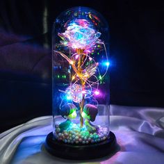 an illuminated flower in a glass dome with beads and lights on the base is sitting on a table