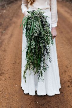 a woman in a long white dress holding a bunch of greenery on the side of a dirt road