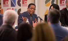 a group of people sitting at a table with microphones in front of them and one man speaking