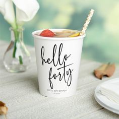 a white cup with a straw and some fruit in it sitting on a table next to flowers