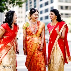 three women in red and gold saris are smiling at each other on the street