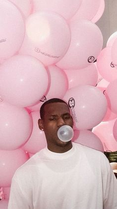 a man standing in front of pink balloons blowing bubbles on his face and nose, while wearing a white t - shirt