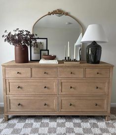 a dresser with flowers and candles on it in front of a mirror, lamp and framed pictures