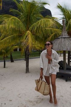 a woman is walking on the beach carrying a straw bag and wearing sunglasses with palm trees in the background