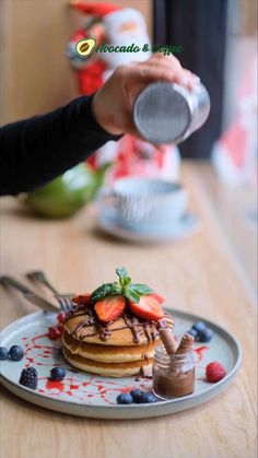 a person is pouring syrup on a stack of pancakes with strawberries and blueberries