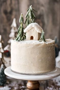 a white frosted cake topped with a small house and pine trees on top of it