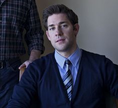 a man wearing a sweater and tie sitting in front of another man with his hand on his hip