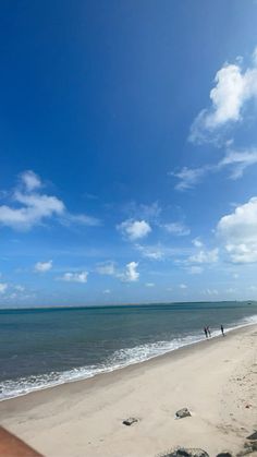 two people are walking on the beach in front of the water and blue sky with white clouds