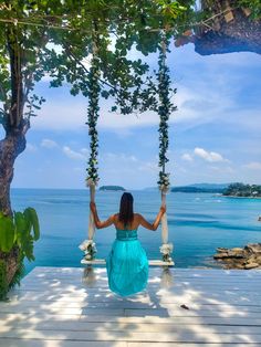 a woman in a blue dress sitting on a porch swing overlooking the ocean and trees