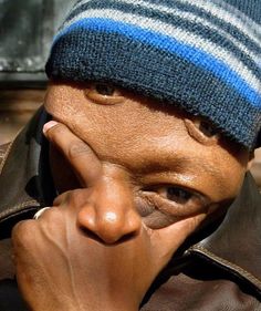 an old man wearing a blue and gray hat covering his face with his hand while looking at the camera