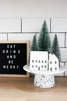 a small white house sitting on top of a cake plate next to a sign that says eat drink and be merry