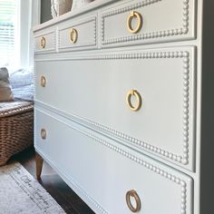 a white dresser with gold handles and knobs in a living room next to a window