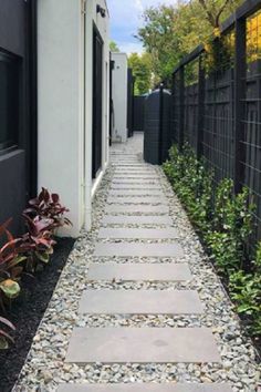 a stone walkway between two buildings with black fence and plants in the foreground on either side