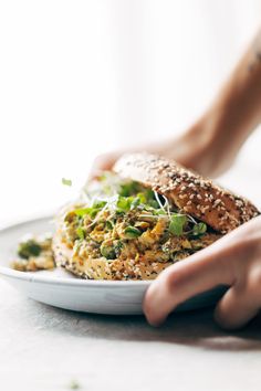 a person is holding a bagel sandwich on a white plate with sprouts