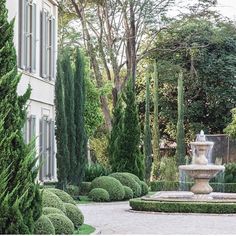 a fountain surrounded by trees and bushes in front of a building with white trimmings