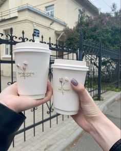 two people holding up coffee cups in front of a fence