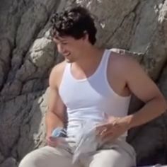 a man in white shirt sitting on rock with cell phone and water bottle next to him