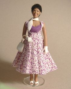 a doll wearing a purple dress and white gloves is standing on a clear base holding a purse