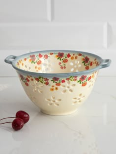 a white bowl with red and yellow flowers on it next to a cherries sprig