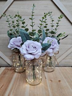 three glass vases filled with purple flowers on top of a wooden table next to a wall