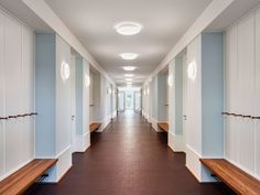 a long hallway with white walls and wooden benches on both sides, along with brown tile flooring