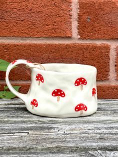 a white cup with red mushrooms on it sitting next to a green leafy plant