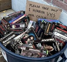 a trash can filled with various types of movies and dvds next to a sign that says please take only 1 horror vhss per child