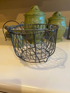 a wire basket sitting on top of a counter next to two green vases with lids