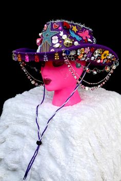 All eyes will be on you in this fabulous purple cowgirl hat covered to the brim with rainbow galore. From the front hang colorful gummy bears and from the sides hang three colorful beaded chains. The hat is covered with stars, hearts, flowers, rhinestones, butterflies, pearls, disco balls, mini animals and treats, and silver tinsel. It has a purple and lace neck tie. Playful Adjustable Hat For Carnival, Playful Adjustable Hats For Carnival, Fun Adjustable Hat For Carnival, Playful Adjustable Carnival Hat, Fun Adjustable Mini Hats For Carnival, Playful Purple Adjustable Hat, Fun Adjustable Hats For Carnival, Funky Adjustable Party Hats, Funky Festival Hats With Curved Brim