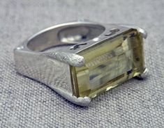 This ring is ornately delicate for a cocktail ring.  It's chunky at the top but tapers towards the bottom to really lighten it up.  It features a pale citrine baguette, prong set at the four corners on the top of the band.  This citrine is the color of sunshine or lemon chiffon and is a gemstone for both the Gemini and Virgo zodiac signs.  The band itself has been gently squared with small round cubic zirconia set into the lower corners on both sides.  This squaring of the band keeps the ring from spinning or twisting on your finger so that the citrine will always stay facing up.  I've done some detailing to the band - adding some decorative carving on the sides just under the citrine and adding a satin texture to the prongs that hold the citrine in place.  Dimensions: size: 7-1/2 citrine: Elegant Sterling Silver Large Stone Topaz Ring, Elegant Citrine Rings With Large Stone, Elegant Sterling Silver Topaz Ring With Large Stone, Elegant Silver Topaz Ring With Large Stone, Gemini And Virgo, Lemon Chiffon, Body Jewelry Piercing, Citrine Ring, Virgo Zodiac