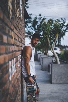 a man leaning against a brick wall holding a skateboard in his hand and looking at the camera