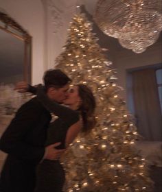 a man and woman kissing in front of a white christmas tree with lights on it