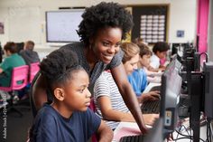 a woman helping a child use a laptop computer in a classroom with other children sitting at desks