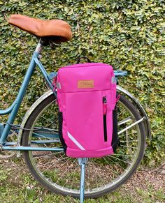 a pink backpack sitting on the back of a blue bike in front of a hedge