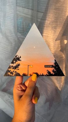 a hand holding up a piece of paper with trees on it and an orange sky in the background