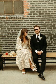 a bride and groom sitting on a bench in front of a brick wall