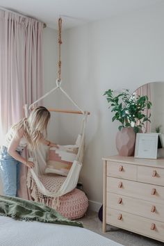 a woman sitting in a hammock chair next to a dresser and window with pink curtains