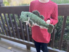 a young boy holding a stuffed alligator toy in his hand on a porch with trees and bushes behind him