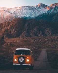 an orange and white van driving down a dirt road in front of snow capped mountains