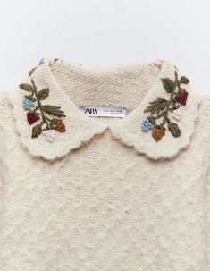 a white sweater with flowers and leaves on the collar is hanging from a hanger